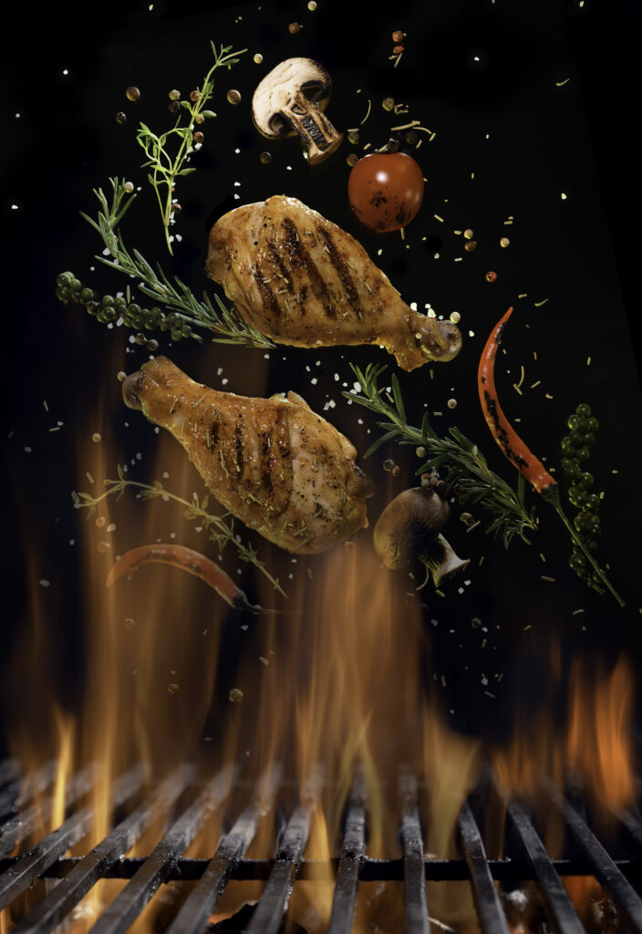 Chicken legs and spicy ingredients flying above flaming red barbeque grill